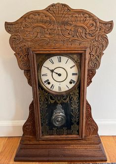 an ornate wooden clock with roman numerals sits on a wood floor in front of a white wall