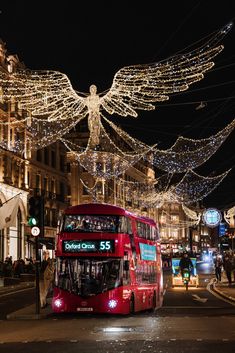 a red double decker bus driving down a street next to tall buildings with christmas lights