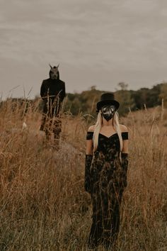 a woman in a black dress and mask walking through tall grass with two other people behind her