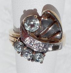 "This great, vintage two-tone ring shows detail and flair typical of the 1940s. The asymmetrical design is composed yellow gold, white gold, and four light blue topaz gemstones. The white gold \"ribbon\" is chased with tiny orange blossoms and decorated with milgrain. It separates the three smaller stones from the one larger stone. The fanned yellow gold section, which shares the side with the larger stone, has two slits and a little curl in the corner. The asymmetry even extends to the shank, w Vintage White Gold Multi-stone Ring, Vintage Multi-stone Diamond Ring Collectible, Vintage Multi-stone Diamond Ring For Collectors, Vintage White Gold Multi-stone Diamond Ring, Vintage Multi-stone Diamond Ring In White Gold, 14k Gold Art Deco Topaz Ring For Anniversary, Vintage 14k Gold Rings With Accent Stones, Vintage White Gold Topaz Ring With Center Stone, Vintage Topaz Ring With Center Stone For Anniversary