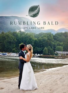 a bride and groom kissing on the beach in front of a lake with mountains behind them