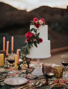 a white wedding cake with red flowers on top and candles in the background at an outdoor dinner table