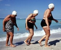 three older women in bathing suits walk along the beach with their backs to the water