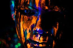 the face of a woman with bright paint all over her body and hands, in front of a black background