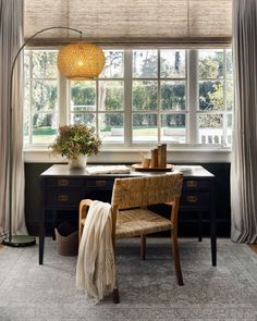 a desk and chair in front of a window with drapes on the windowsill