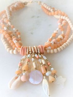 "20\" Carved Natural Wood, Mother of Pearl, Peach Beads & Peach CoCo Shell 4 Strand Necklace with Bead Dangles Brass Hook Clasp This 4 Strand Necklace is Hand Strung with Natural Wooden Beads, Peach Wooden Beads, Mother of Pearl Beads and Peach Beads The Hand Carved Wooden Sea Shell Pendant Hangs in Front by Rings of Small Wooden Beads on Top of Peach CoCo Beads There are 6 Strands of Beads Dangling on Top of the Wooden Sea Shell with a Carved Mother of Pearl Starfish & Natural Shells on Orange Multi-strand Jewelry For Beach, Orange Multi-strand Jewelry For The Beach, Peach Colored Jewelry With Colorful Round Beads, Orange Multi-strand Beaded Necklaces For Beach, Peach Beaded Jewelry With Round Beads, Apricot Beaded Round Bead Jewelry, Bohemian Peach Round Beads Jewelry, Pink Beaded Bohemian Shell Necklace, Pink Bohemian Beaded Shell Necklace