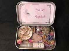 an open tin with various items inside on a black tableclothed surface and the words you're magickal written in pink