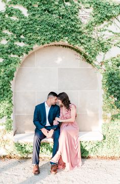 a man and woman sitting on a bench in front of a wall covered with ivy