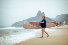 a woman walking on the beach with a surfboard in her hand and mountains in the background