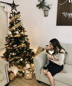 a woman sitting on a couch holding a baby next to a christmas tree