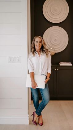 a woman standing in front of a white wall holding a folder and posing for the camera