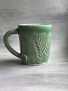 a green coffee mug with leaves on the side and a button in the middle, sitting on a table