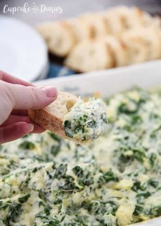 a hand holding a piece of bread with spinach and cheese on it in a casserole dish