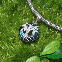 a blue and silver necklace with leaves on it sitting in the grass next to a leafy green plant