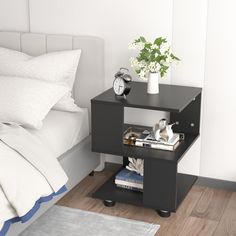 a nightstand with books, magazines and a clock on it next to a white bed