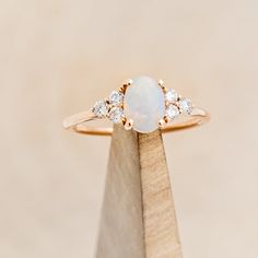 a white opal and diamond ring sitting on top of a wooden stand in front of a beige background