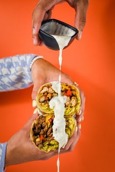 a person pouring sauce onto a burrito on an orange background while holding a cell phone in their hand