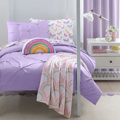 a bed with purple sheets and pillows in a bedroom next to a white nightstand table