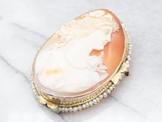 "This cameo features wonderful, crisp details, and a great contrast between the creamy white carving and the soft russet of the background. A string of seed pearls pulls the piece together, drawing on the color of the profile and the floral details of the gold. Metal: 14K Yellow Gold Gem: Shell Cameo Gem Measurements: 34.4 x 45.6 mm, Oval Accents: Seed Pearls Measurements: 40 x 55 mm Marks: \"X\" Stamped To view a video of this piece check out the link below: https://vimeo.com/802860432 SKU #: A Exquisite White Brooch For Formal Occasions, Elegant Formal Brooch With Intaglio Detail, Elegant Intaglio Brooch For Formal Occasions, White Cameo Brooch For Formal Occasions, White Cameo Brooches For Formal Occasion, Luxury Cameo Brooches, Luxury Cameo Brooch For Wedding, Luxury Intaglio Brooch For Formal Occasions, Heirloom White Brooch For Formal Occasions