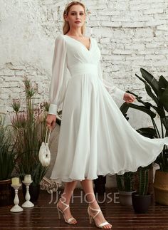 a woman standing in front of a brick wall wearing a white dress with long sleeves