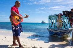 a man playing guitar on the beach while people watch from the water behind him with boats in the background