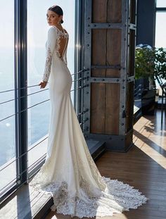 a woman standing in front of a window wearing a long sleeved wedding dress with an open back