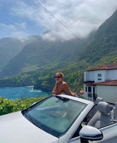 a woman sitting on top of a car next to a body of water with mountains in the background