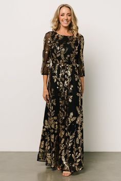 a woman wearing a black and gold floral print dress with sheer sleeves, smiling at the camera