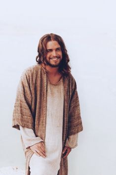 a man with long hair and a beard standing on the beach wearing a brown cardigan