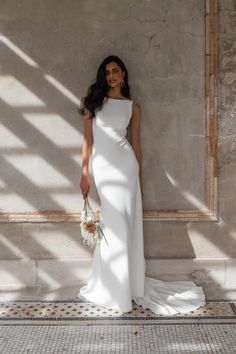 a woman standing in front of a wall wearing a white dress and holding a bouquet