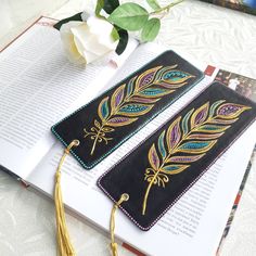 two bookmarks with gold and blue feathers on them sitting next to an open book