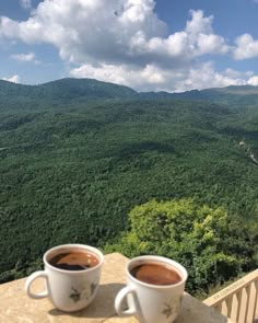 two cups of coffee sitting on top of a table next to each other with mountains in the background