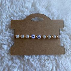 a white and gold beaded name bracelet on top of a brown card with the word ao