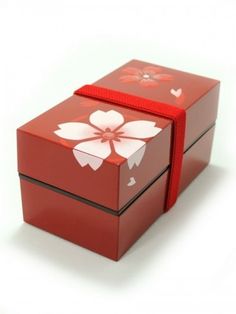 an open red box with white flowers on the lid and ribbon around the top, sitting on a white surface