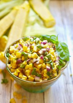 a green bowl filled with corn salad on top of a wooden table next to sliced vegetables