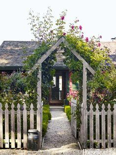 a white picket fence with flowers growing over it and a house in the background,