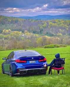 a person sitting on a bench next to a blue car in a field with mountains in the background