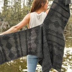 a woman holding up a black shawl in front of a body of water with trees in the background