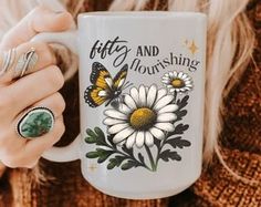 a woman is holding a coffee mug with daisies and butterflies on it, which reads fifty and flourishing