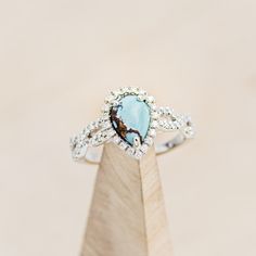 a diamond and turquoise stone ring on top of a wooden stand in front of a beige background