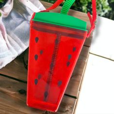 a plastic watermelon cup with a green lid on a wooden table next to a towel