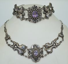 The Demi Parure is from the Netherlands, it dates from the 20/30s. The set consists of a necklace as well as a matching bracelet. The suite is crafted from sterling silver with earth mined bezel set amethysts. The bracelet is rather chunky with the center spanning a length of 1.25 Inches. The bracelet features 5 semi curved links that are hollow in construction. The design is raised with a rich patina. The clasp is a box clasp with a safety. The necklace features a similar design with a bit of variation. The necklace features a small box clasp with an eye safety. Both pieces are in excellent condition with a rich patina and normal wear. This is a very well crafted set. They are hallmarked with an I dating this piece to be essayed from 1906-1953. Measurements: Necklace: 17.25 inches in leng Elegant Amethyst Jewelry With Sterling Silver Clasp, Elegant Sterling Silver Amethyst Bracelet For Formal Occasions, Antique Formal Jewelry With Sterling Silver Clasp, Antique Jewelry With Sterling Silver Clasp For Formal Occasions, Art Deco Silver Amethyst Jewelry, Art Deco Amethyst Silver Jewelry, Silver Amethyst Art Deco Jewelry, Vintage Amethyst Jewelry With Polished Finish, Silver Art Deco Jewelry