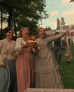 three women dressed in period clothing holding a cake and waving at the camera with their arms wide open