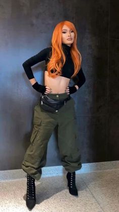 a woman with long red hair standing in front of a wall wearing green pants and black boots