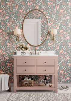 a bathroom with pink vanity and floral wallpaper on the walls, along with a round mirror