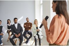 a woman holding a microphone in front of a group of men and women sitting on chairs