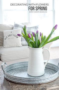 a white vase with purple flowers in it on a tray near a couch and window