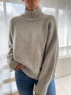 a woman standing in front of a window wearing a gray sweater