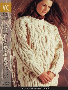 a woman wearing a white cabled sweater with her hands on her hips, standing in front of a tree