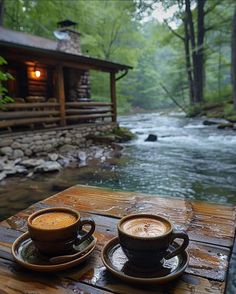 two cups of coffee sitting on top of a wooden table next to a river in the woods